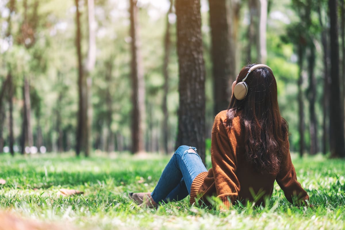 Joven sentada en el pasto en un bosque, escuchando música con auriculares.