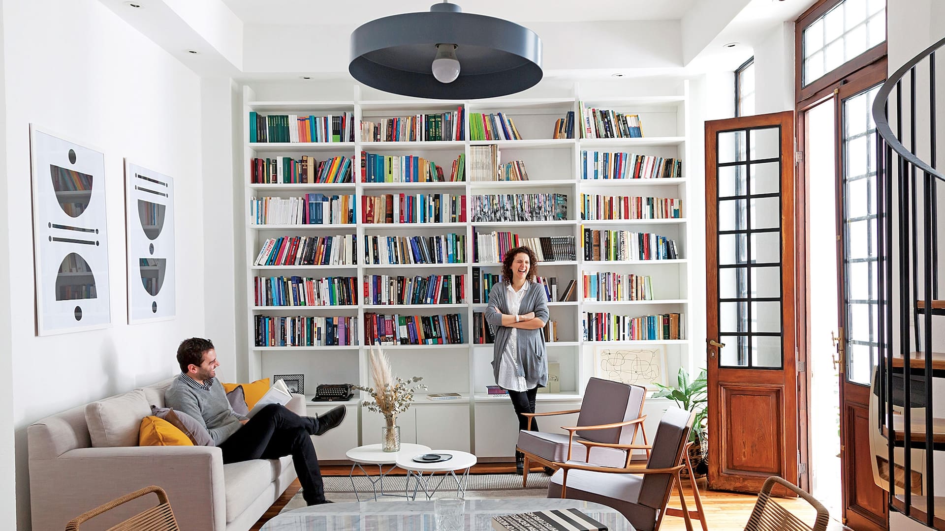 Joy y su marido en el living equipado con un amplio sofá (Loft) y dúo de sillones individuales heredados. La biblioteca (Melazza) es un diseño de Ideï. Los cuadros también son obra de ella.