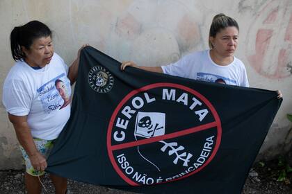Kelly Christina da Silva, a la derecha, cuyo hijo de 23 años murió a causa de las heridas provocadas por los cortes de un hilo de cometa recubierto de fragmentos afilados, sostiene junto a su madre Maria Enilda da Silva una bandera en la que se lee en portugués: "El hilo cortante mata. Esto no es una broma", en Río de Janeiro, el lunes 29 de julio de 2024. (AP Foto/Bruna Prado)
