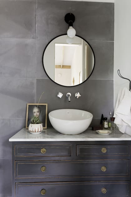 La cómoda fue reciclada con pátina gris oscuro (Taller Alsina). Sobre la mesada de baño en mármol de Carrara, bacha ‘Bol’ (Roca). Espejo circular (Mercado Libre). Aplique de luz ‘James’ negro mate (Pura Iluminación). Porcelanato oscuro (Ilva). Grifería ‘Alerce’ (FV).