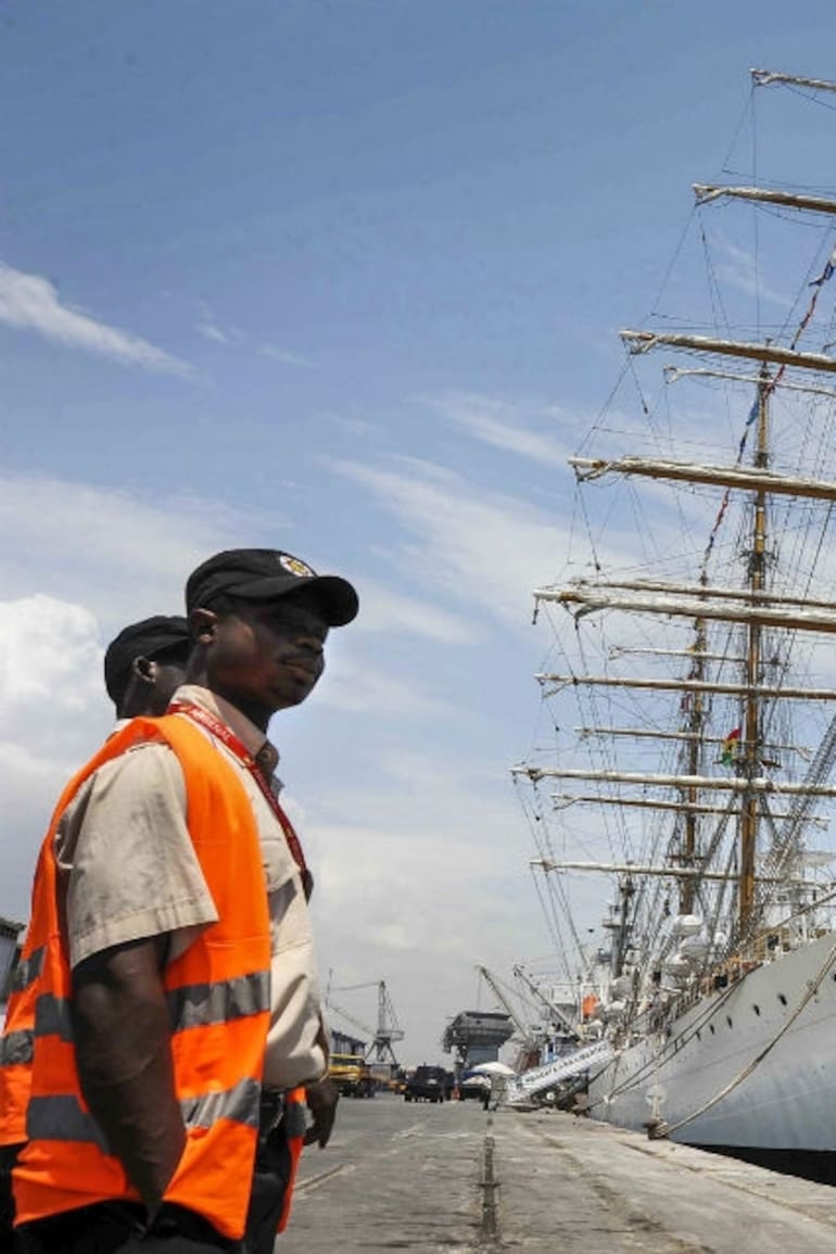 El buque escuela, en el puerto de Tema, cerca de Accra, la capital del país africano, con la bandera local. A la izquierda, dos funcionarios locales que trabajaban en el muelle.