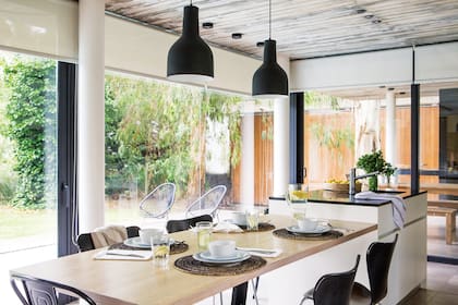 La mesa de la cocina está enchapada en roble claro y tiene base de hierro pintado a fuego (Metro Maderas). Arriba, dos lámparas colgantes (Casa Blanco Iluminación) demarcan el espacio del comedor con su haz de luz. Sillas de madera y una manta en uno de los respaldos (Elementos Argentinos).