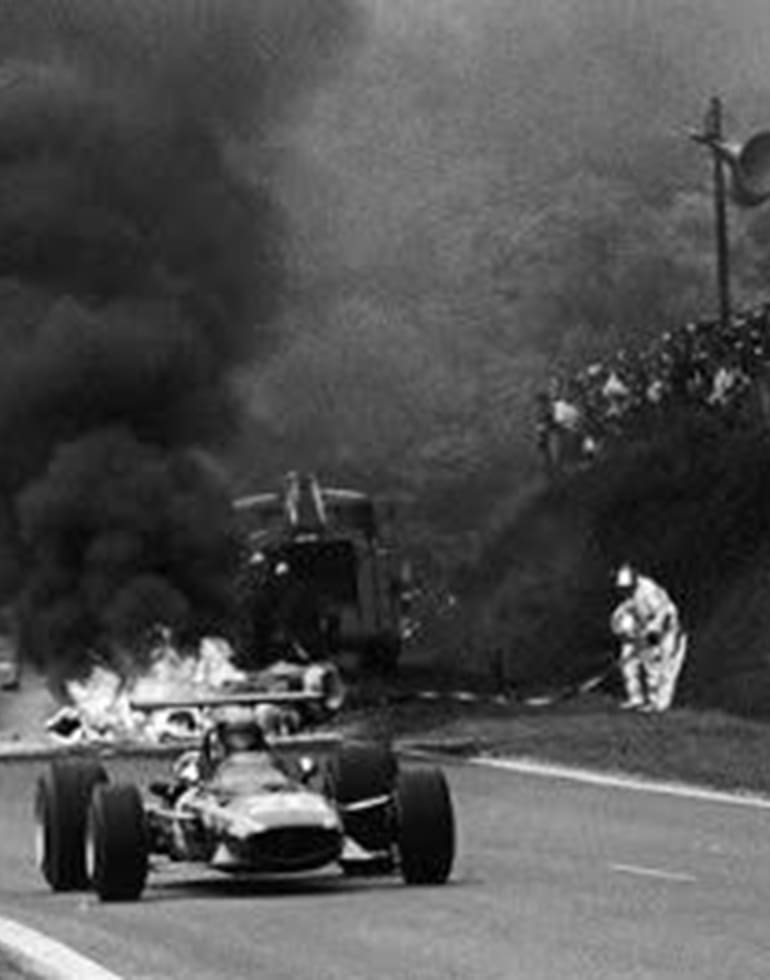 Los autos pasan por la zona del accidente, donde el auto de Jo Schlesser se consume en medio del fuego