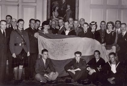 Los voluntarios sobrevivientes del buque Andalucía Star convertidos en voluntarios posan con el embajador Dr. Miguel Ángel Cárcano (centro de la imagen). John Easdale, oriundo de Jujuy instructor de vuelo en la RAF, sostiene la bandera Argentina que fue rescatada del buque antes que se hundiera. (Archivo John Linton Easdale).