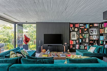 El fondo de la biblioteca tiene el mismo revestimiento que la pared donde se apoya el televisor. Hecho a medida, más profundo que lo habitual, el sillón se tapizó en terciopelo y se lo combinó con almohadones de motivos botánicos.