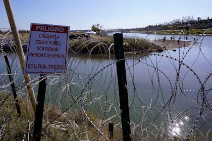Migrantes cruzan el río Bravo a Estados Unidos desde México detrás de un hilo de cuchillas y un cartel que advierte que cruzar es peligroso e ilegal, el miércoles 3 de enero de 2024