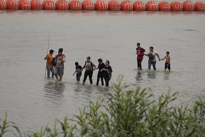 Migrantes que cruzaron el Río Grande (Bravo) desde México caminan frente a enormes boyas colocadas como una barrera fronteriza flotante en el río, el miércoles 12 de julio de 2023, en Eagles Pass, Texas
