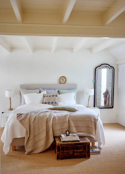 Moquete de fibras naturales, velador con base de tronco, baúl vintage, pie de cama y uno de los almohadones tejidos a mano: detalles que le dan un sofisticado toque rústico a un cuarto blanco.