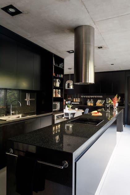 Muebles de madera laqueados en negro satinado (Villanueva-Cseh) hechos a medida con accesorios cromados. Techo de hormigón con luminarias embutidas; allí también se acopla la campana cilíndrica de aluminio. Mesada, alzada e isla comparten el granito negro pulido.