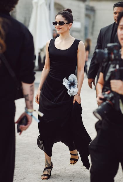 Para el desfile de Dior, Rosalía optó por un sencillo vestido negro sin mangas, con escote redondo amplio y tirantes anchos, ceñido en el pecho y muy fluído en su caída. ¿El detalle? Una gran flor de tela aplicada en la cintura, que permite un plisado lateral de la falda. La cantante completó su outfit con sandalias negras y lentes de sol