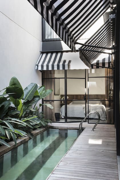 Para preservar las ventanas, se instalaron toldos con el mismo patrón a rayas de la pileta y los espacios comunes del edificio. El dormitorio está separado por cerramientos de vidrio repartido y cortinados de terciopelo en tono habano (Maia Grandoli).