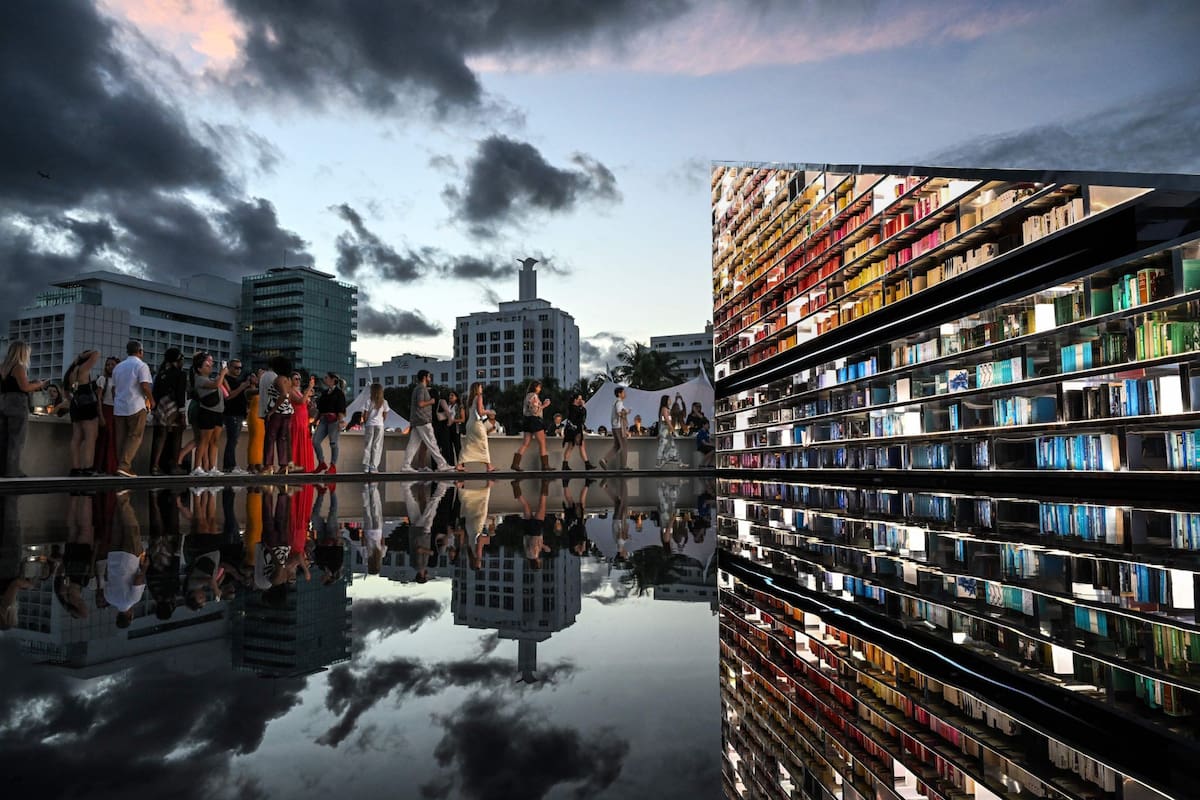 People look at the exhibition "LIBRARY OF US" by British artist Es Devlin, a 50-foot-wide rotating triangular bookshelf containing 2,500 books that have shaped the artist's life and practice, during Art Basel 2025, in Miami Beach, Florida, December 2, 2025. (Photo by CHANDAN KHANNA / AFP) / RESTRICTED TO EDITORIAL USE - MANDATORY MENTION OF THE ARTIST UPON PUBLICATION - TO ILLUSTRATE THE EVENT AS SPECIFIED IN THE CAPTION