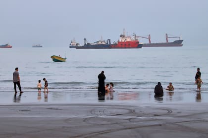Personas pasan el tiempo en una playa mientras varios buques comerciales permanecen fondeados en el estrecho de Ormuz, frente a Bandar Abbas, Irán