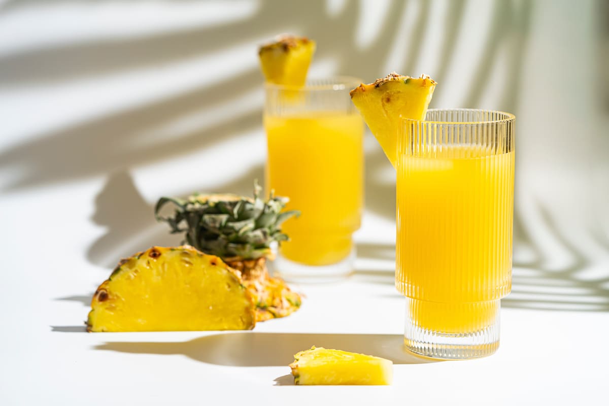 Pineapple cocktail or juice in two glasses with ice on white background with palm leaves shadows