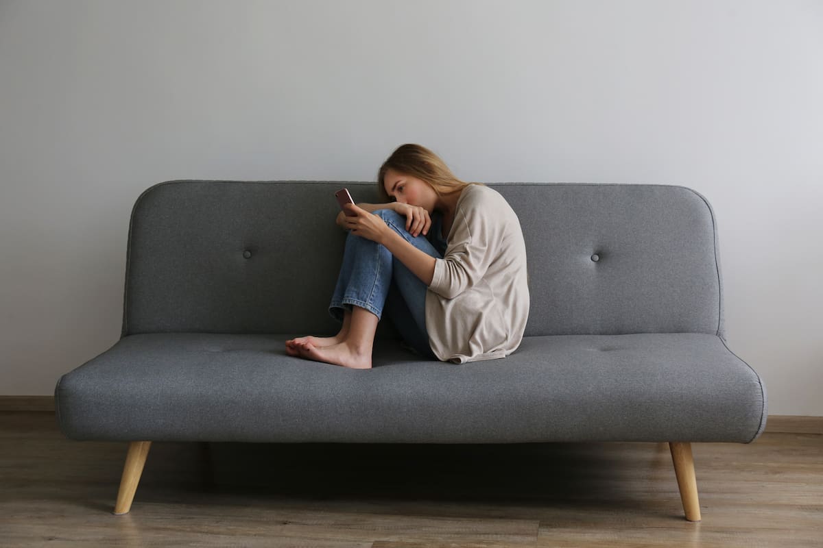 Portrait of beautiful young woman with depressed facial expression sitting on grey textile couch holding her phone. Cyber bullying victim concept. Sad female in her room. Background, copy space.