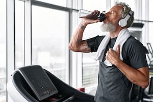 Portrait of mature grey-haired sportsman, drinking water from bottle after workout. Handsome bearded eldrely man with white towel on shoulders drinking to stay hydrated at gym.