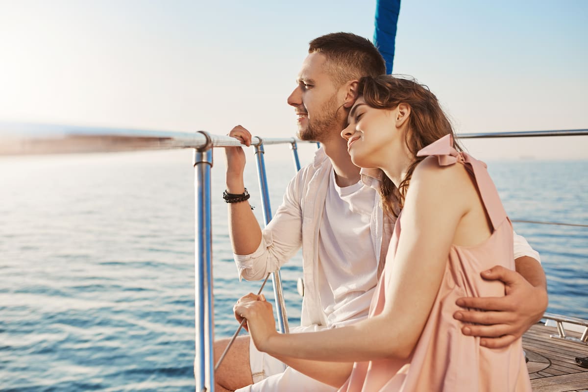 Portrait of stylish couple in love, hugging while sitting on bow of private yacht and enjoying view of sea. Husband took his wife to beautiful warm country, celebrating honeymoon. Vacation and travel concept.