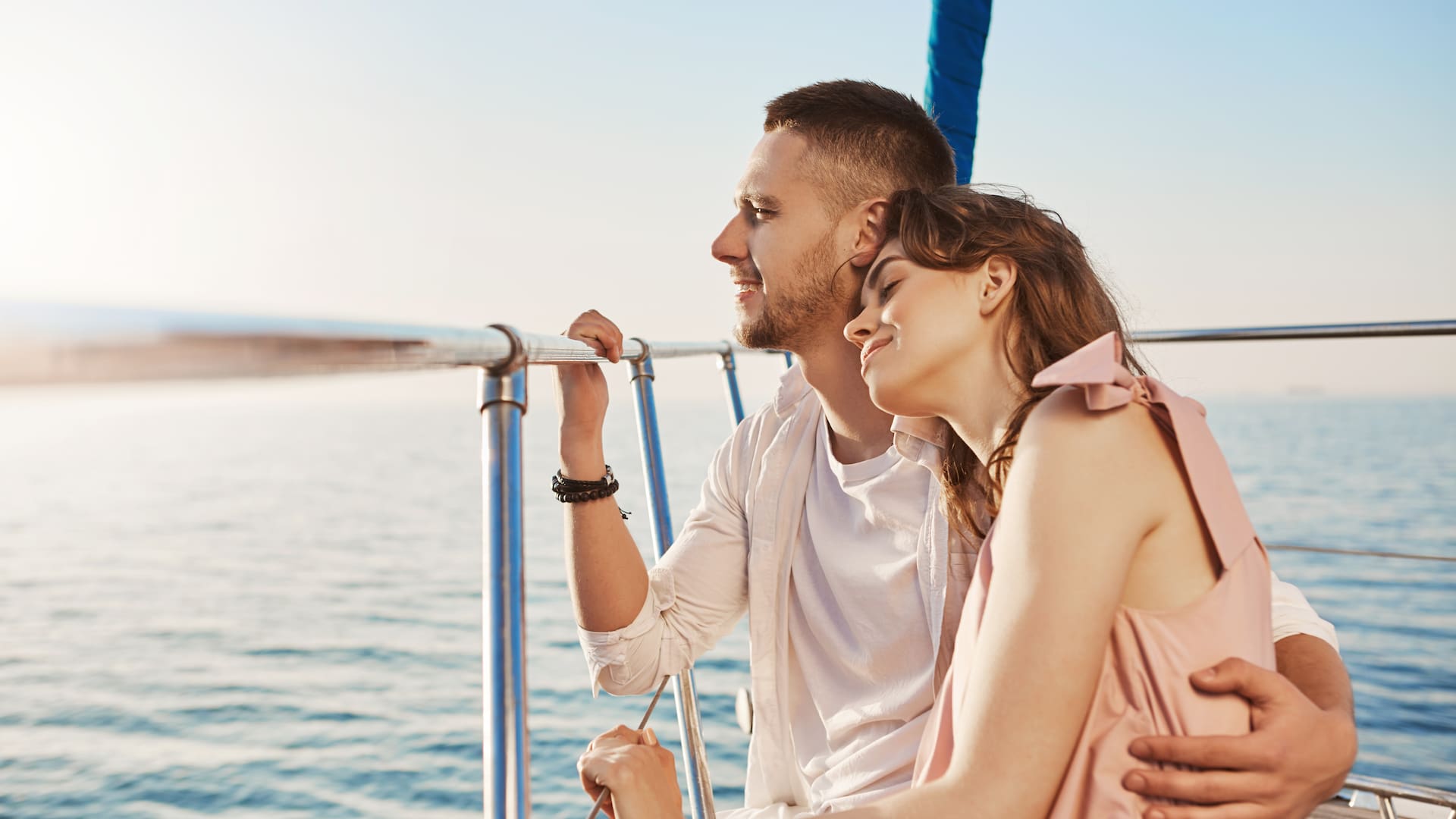 Portrait of stylish couple in love, hugging while sitting on bow of private yacht and enjoying view of sea. Husband took his wife to beautiful warm country, celebrating honeymoon. Vacation and travel concept.