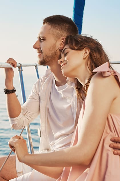 Portrait of stylish couple in love, hugging while sitting on bow of private yacht and enjoying view of sea. Husband took his wife to beautiful warm country, celebrating honeymoon. Vacation and travel concept.