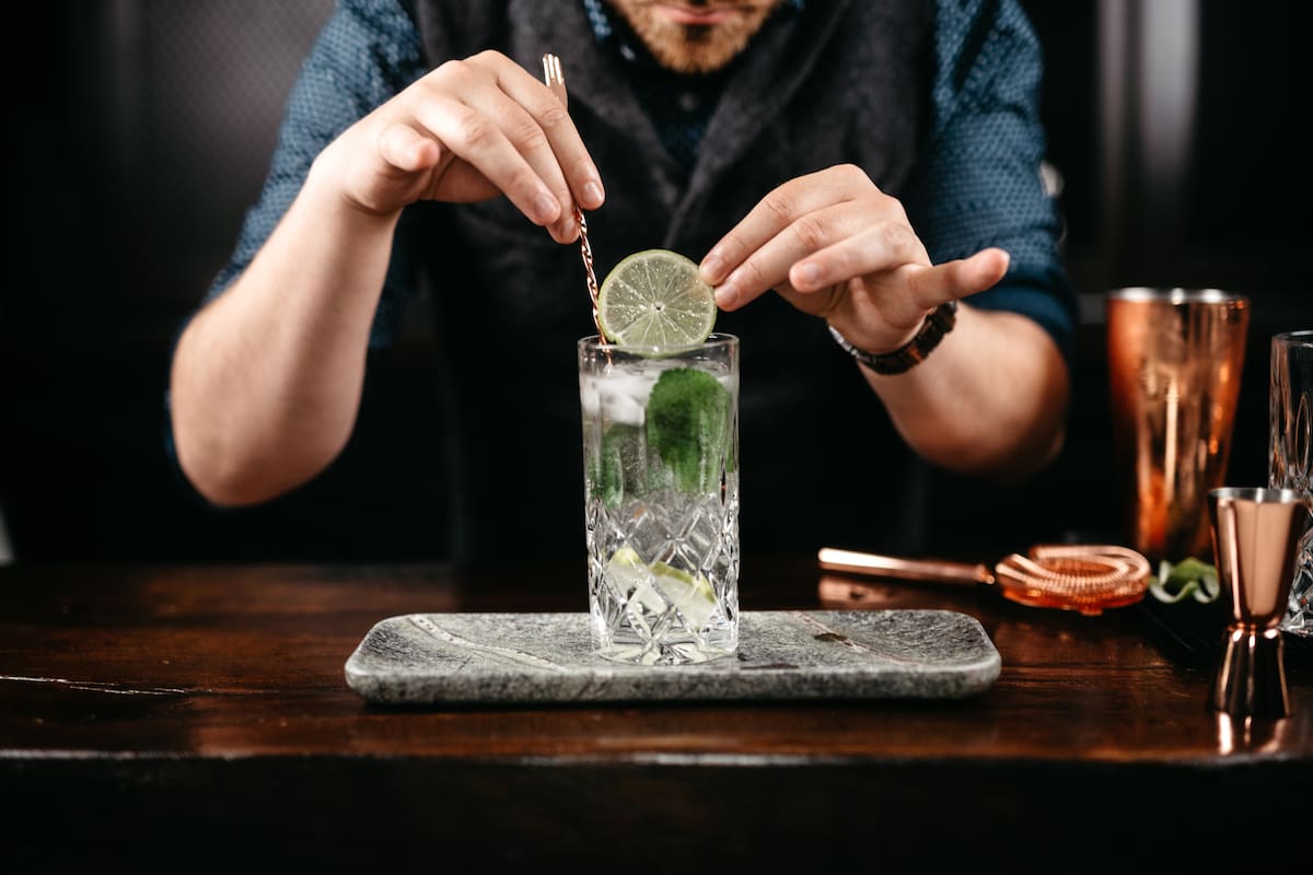 Professional bartender pouring and preparing gin and tonic with lime at bar counter. Details of mixology