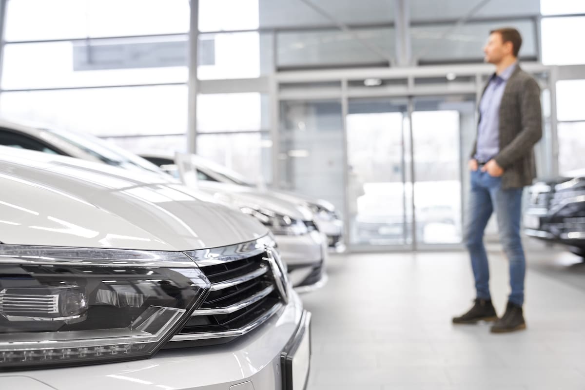 Selective focus of headlights of big white car in auto salon. Young male customer looking at new cars and choosing standing at background inn car showroom. Concept of buying automobile.