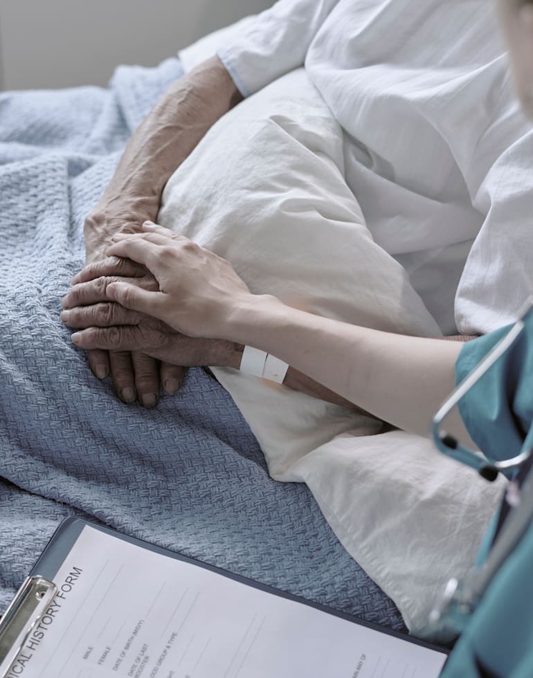Senior Caucasian patient lying in hospital bed receiving comforting touch from young adult Caucasian female nurse, medical form and stethoscope visible in clinical setting