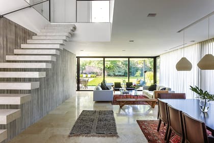 Siguiendo el tabique de hormigón visto, se accede al espacio que integra comedor y living. Alfombra marroquí (Kasbah). Banco V en madera (Sticotti). Sillones restaurados, dúo de sofás y mesas bajas de mármol ‘Negro Marquina’ y Desert Brown (todo de Estudio MMS). Florero y arreglo (Regiacasa)