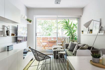 Sillón de tres cuerpos con ‘puf ’, tapizado con pana ‘Velvet’ gris (De Levie). Dúo de mesas ‘nido’ triangulares de hierro con tapa de madera laqueada. Lámpara de pie blanca (Kolor). Alfombra gris con rayas (Rugit). Sillón ‘Gaviota’ negro (Increa). Al fondo, el balcón, con mesa y sillas de madera.