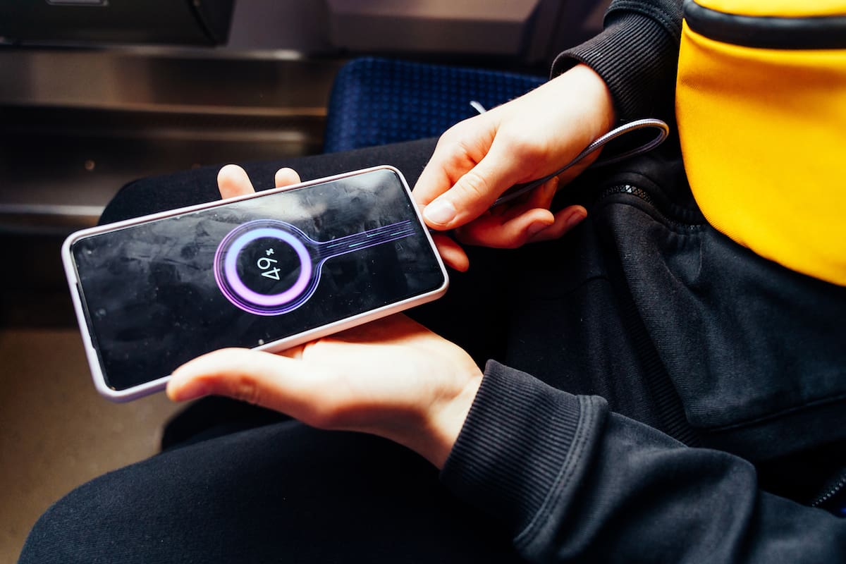 Smartphone charging in teenager's hands, phone shows battery percentage, seated on train in dark clothes and yellow bag, close-up detail of tech usage in public transport