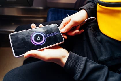 Smartphone charging in teenager's hands, phone shows battery percentage, seated on train in dark clothes and yellow bag, close-up detail of tech usage in public transport