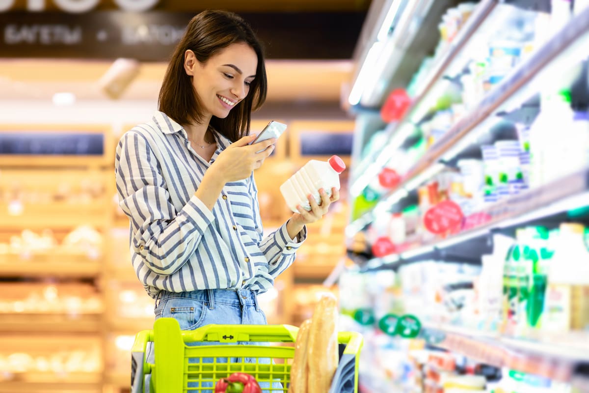 Smiling young woman taking dairy products from shelf in the supermarket, holding bottle and smartphone, scanning bar code on product through mobile phone, walking with trolley cart