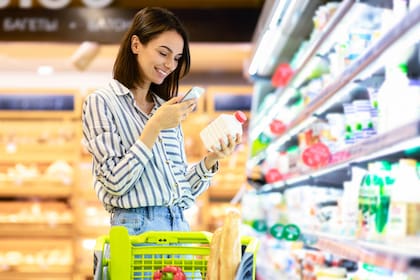 Smiling young woman taking dairy products from shelf in the supermarket, holding bottle and smartphone, scanning bar code on product through mobile phone, walking with trolley cart