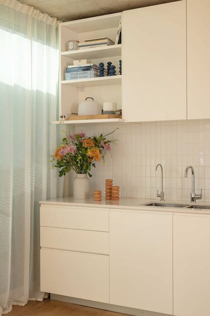 Sobre el fondo neutro, un florero con flores frescas, libros de arquitectura y pequeños objetos en azul y madera adquieren protagonismo, sumando calidez a un espacio concebido desde la sutileza.