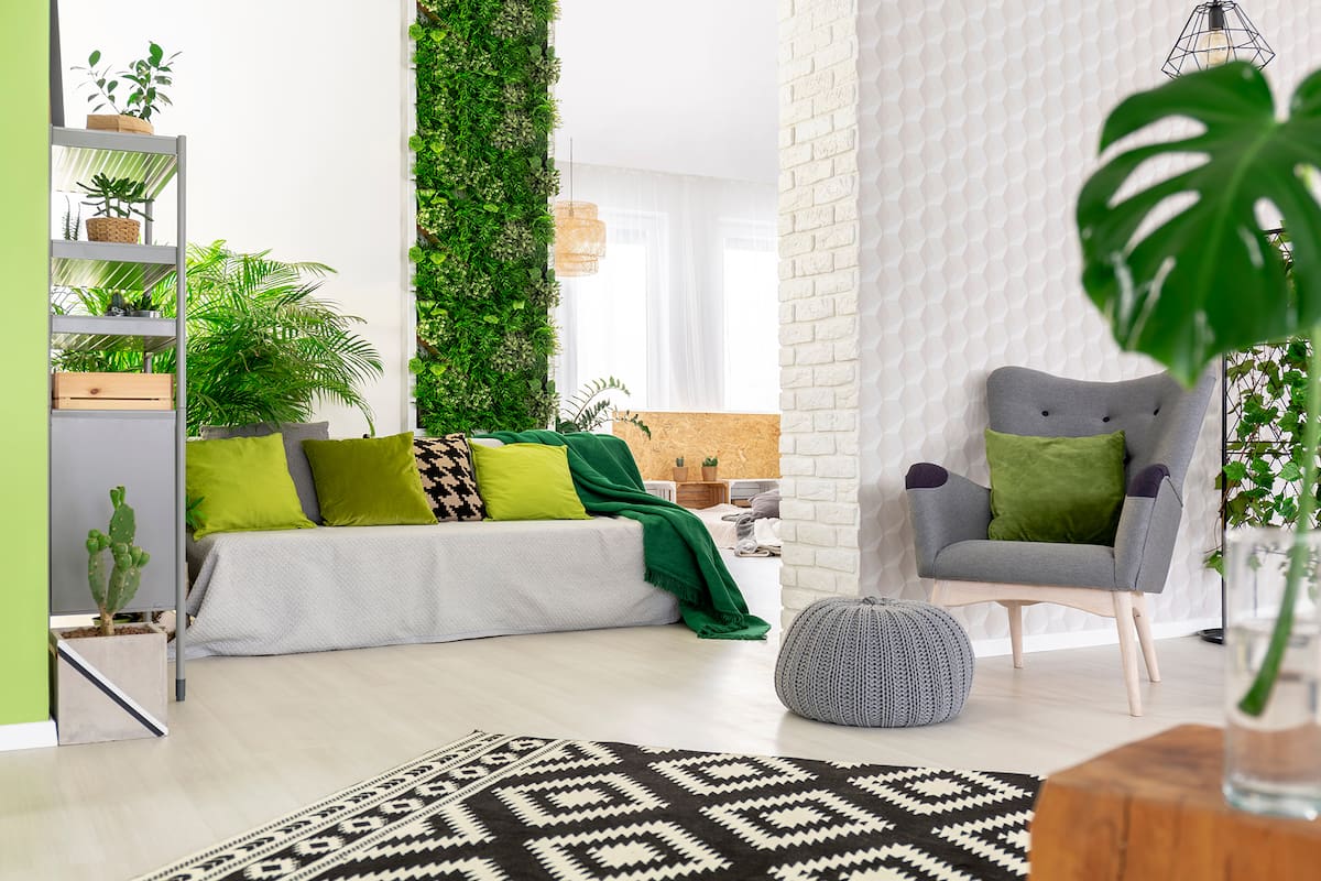 Sofá con almohadas verdes y manta de pie en el espacio abierto salón interior con sillón gris y reposapiés, moqueta en blanco y negro, plantas frescas y un jardín vertical en el fondo