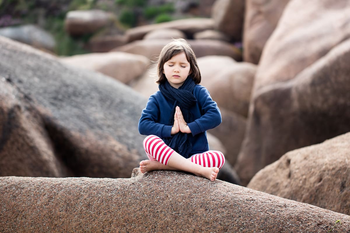 spiritual young kid with eyes closed trying to relax, pray and breathe alone for yoga outdoor, seeking for mindfulness, sitting on giant granite stones, springtime