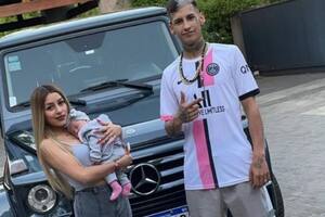 Tamara Báez y L-Gante posan con su pequeña hija, junto a uno de sus vehículos detrás.