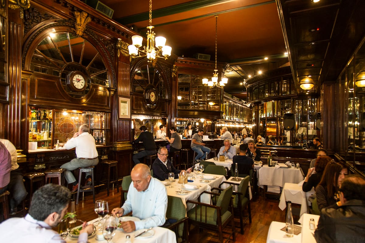 The New Brighton impacta con la barra de madera rojiza y los taburetes donde sentarse a beber un Negroni, la boiserie de cedro en las paredes, el pulido del parqué, los espejos y los biselados, los grandes carteles de bodegas tradicionales que recorren el salón, los vitrales de vidrios, el piano imponente