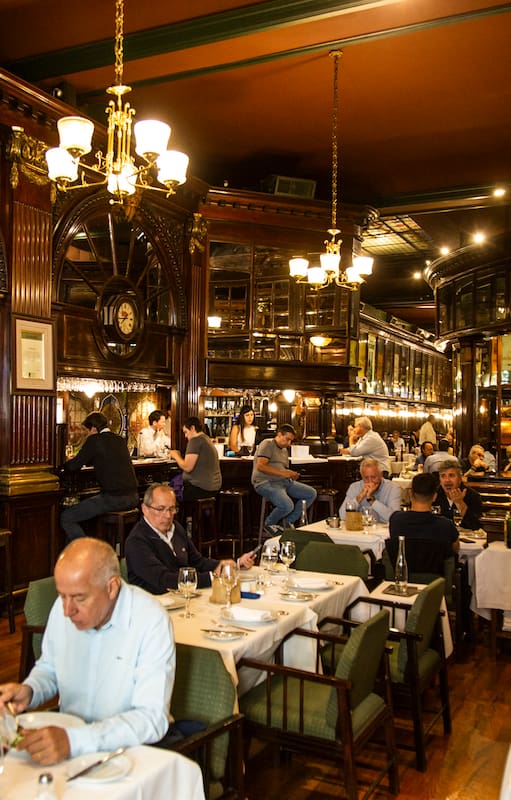 The New Brighton impacta con la barra de madera rojiza y los taburetes donde sentarse a beber un Negroni, la boiserie de cedro en las paredes, el pulido del parqué, los espejos y los biselados, los grandes carteles de bodegas tradicionales que recorren el salón, los vitrales de vidrios, el piano imponente
