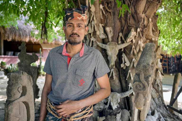 This photo taken on June 24, 2026 shows marapu priest Umbu Remi Deta, who is a maramba (nobleman), posing by a large tree mounted with skulls and jawbones of sacrificial animals alongside traditional stone carvings at Kampung Raja Prailiu at Waingapu in East Sumba, East Nusa Tenggara. Two hours from the tourist beaches of Bali, slavery is alive and well on the Indonesian island of Sumba, where people are still being born into bondage. Eight of out 10 of Sumba's population of 685,000 people are Christian but its older "marapu" belief system, that blends animism and ancestor worship, has deep roots. It divides society into three castes, "the nobles, called 'maramba' -- often large landowners -- ordinary people called 'kabihu' and the slaves/servants, the atas," said rights activist Martha Hebi, author of several studies on the subject. (Photo by BAY ISMOYO / AFP) / To go with Indonesia-Sumba-slavery, SPECIAL REPORT by Eric Bernaudeau