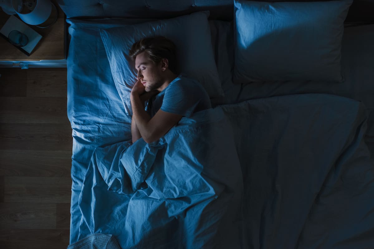 Top View of Handsome Young Man Sleeping Cozily on a Bed in His Bedroom at Night. Blue Nightly Colors with Cold Weak Lamppost Light Shining Through the Window.