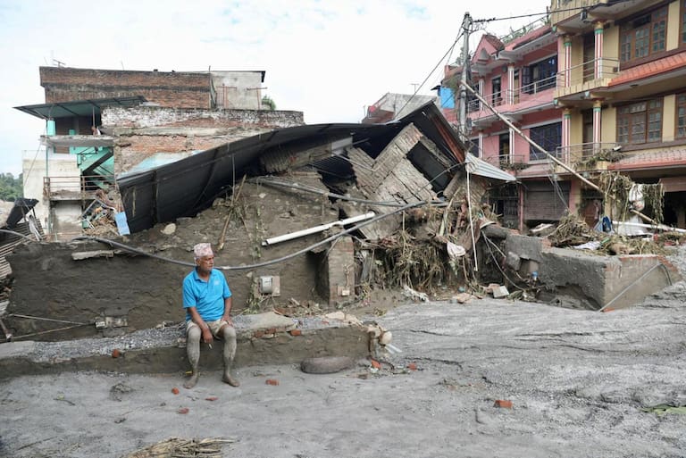 TOPSHOT - A local resident sits near a collapsed house lying deluged in sludge after flash floods at Devighat, Bidur Municipality in Nepal's Nuwakot district on August 27, 2026. Nepal's disaster agency said that 823 people remained out of contact on August 27, more than 24 hours after a deadly flood tore through the border region with Tibet. (Photo by Prabin RANABHAT / AFP)