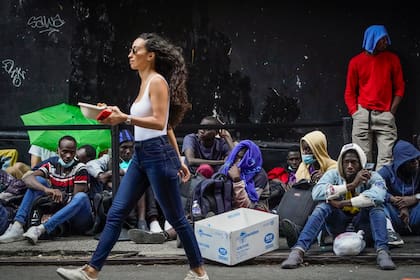Transeúntes caminan frente a migrantes que esperan afuera del Hotel Roosevelt, utilizado por la ciudad de Nueva York como albergue temporal (AP Foto/John Minchillo)
