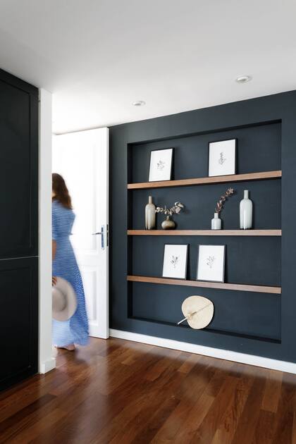 Tres largos estantes a modo de pequeña biblioteca con diferentes objetos decorativos como jarrones con hojas secas y pintada en un tono oscuro para que llame la atención de quien pase por ese espacio.