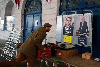 Un agricultor prepara su puesto en el mercado de Salies de Bearn, en el suroeste de Francia, ante dos carteles de campaña del presidente de Francia y candidato centrista a la reelección, Emmanuel Macron, y la candidata de ultraderecha Marine Le Pen, a la derecha, el sábado, el 23 de abril de 2022.  (AP Foto/Bob Edme)