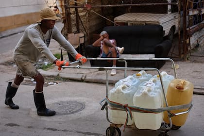Un hombre empuja un carrito lleno de bidones de agua mientras otro descansa en un sofá en una calle de La Habana, Cuba, el jueves 27 de agosto de 2026.