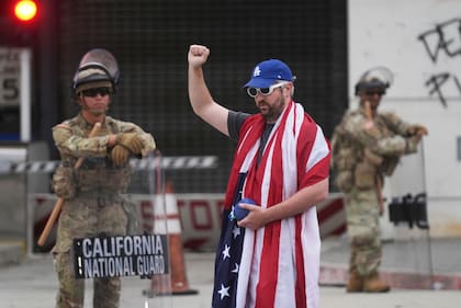Un manifestante cubierto con la bandera de Estados Unidos alza un brazo frente al Centro de Detención Metropolitano y miembros de la Guardia Nacional de California (AP Foto/Damian Dovarganes)