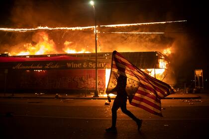Un manifestante lleva una bandera estadounidense invertida, señal de estar en peligro, frente a un edificio en llamas, 28 de mayo de 1010 en Minneapolis. Es parte de una serie de fotos de The Associated Press que ganó el Premio Pulitzer 2021 a la fotografía de noticias de último momento. (AP Foto/Julio Cortez)