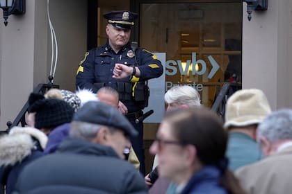 Un oficial de policía de Wellesley, Massachusetts, mira su reloj mientras los clientes y transeúntes forman una fila frente a una sucursal de Silicon Valley Bank, el lunes 13 de marzo de 2023. (AP/Steven Senne)
