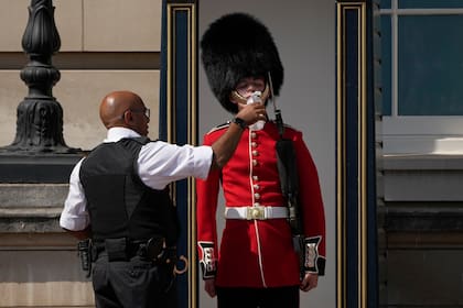 Un oficial de policía le da agua a un soldado británico que lleva un sombrero de piel de oso tradicional, de guardia frente al Palacio de Buckingham, durante la ola de calor en Londres, el lunes 18 de julio de 2022