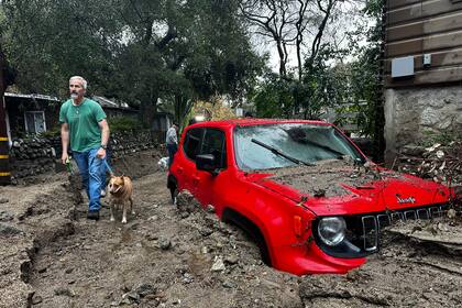 Un residente y su perro caminan junto a un vehículo parcialmente sumergido en lodo tras una tormenta, el viernes 14 de febrero de 2025, en Sierra Madre, California